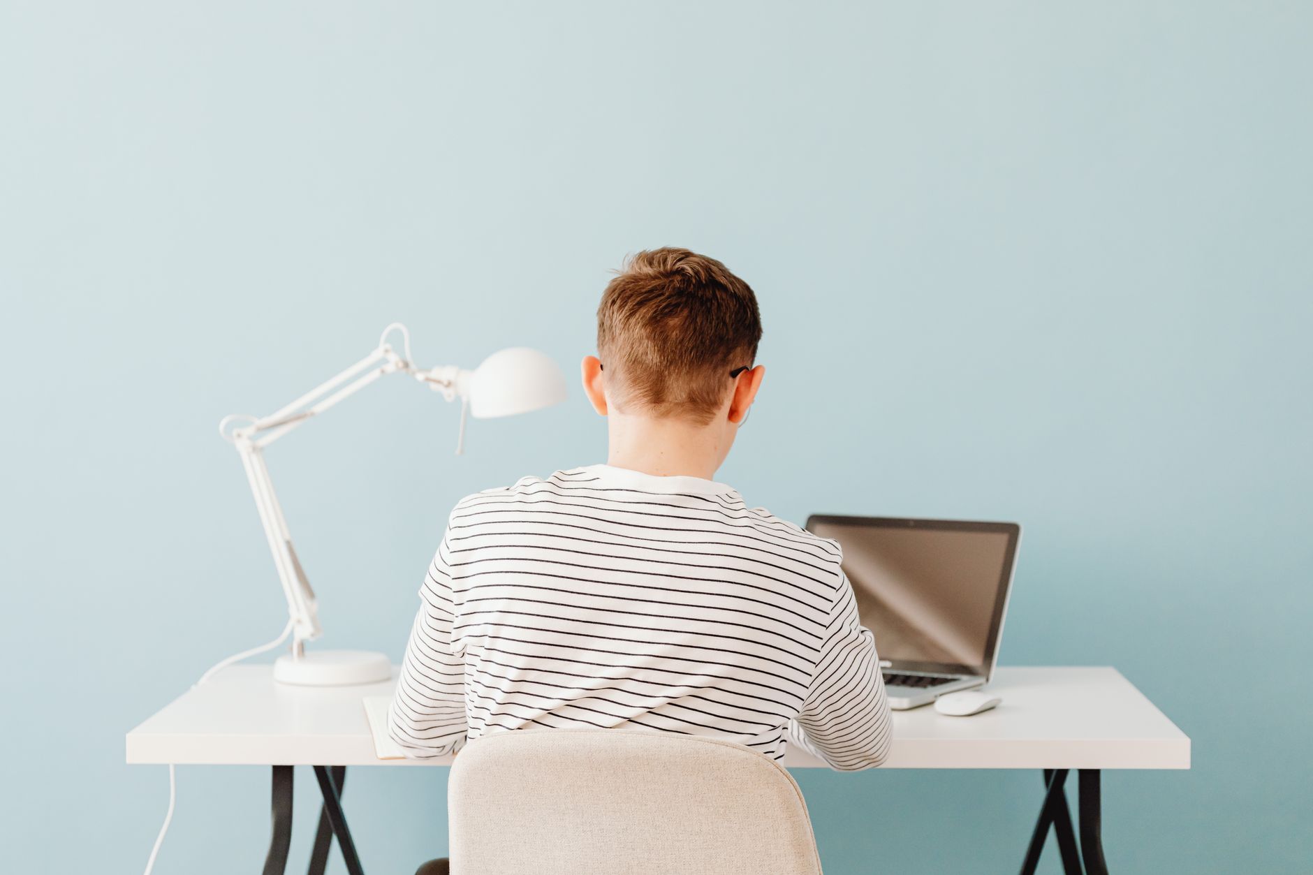 Eine Person im gestreiften Pullover sitzt von hinten gesehen an einem weissen Schreibtisch mit Laptop und weisser Lampe vor einer hellblauen Wand.