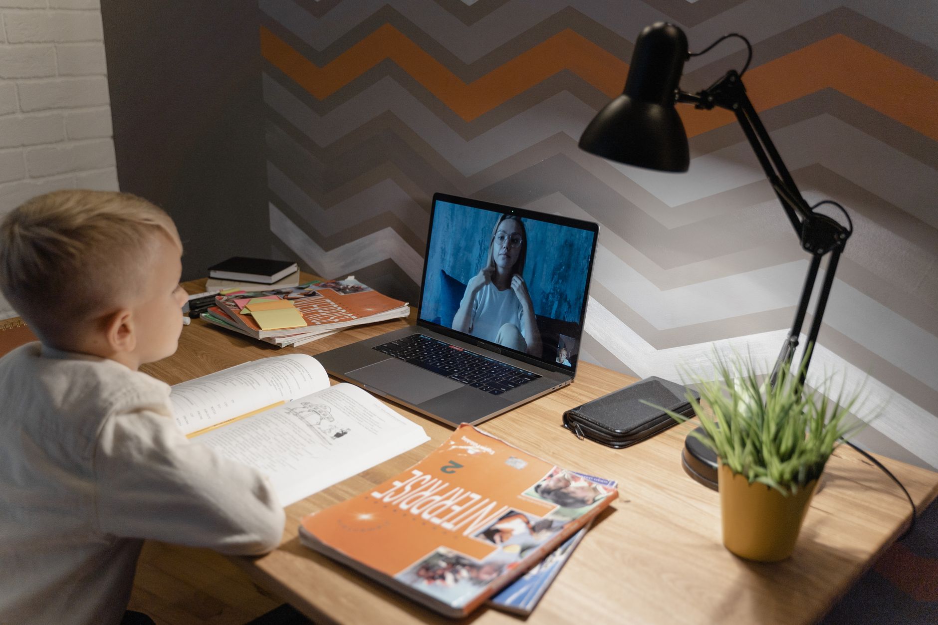 Ein Kind mit blondem Haar sitzt abends an einem Holzschreibtisch mit Schreibtischlampe und blickt auf einen Laptop, auf dem ein Videoanruf läuft.