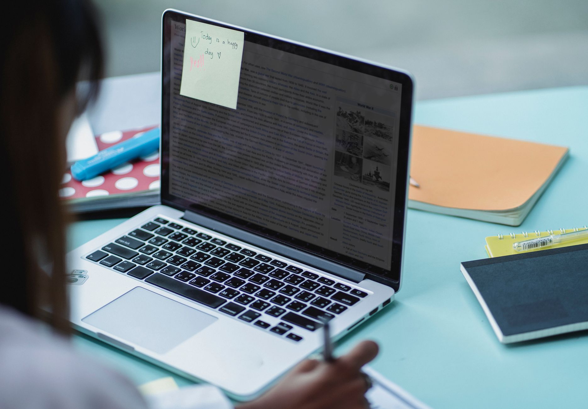 Blick über die Schulter einer Person auf einen aufgeklappten Laptop, auf dem Bildschirm klebt ein Notizzettel; daneben liegen Notizbücher und ein blauer Textmarker auf einem hellblauen Tisch.