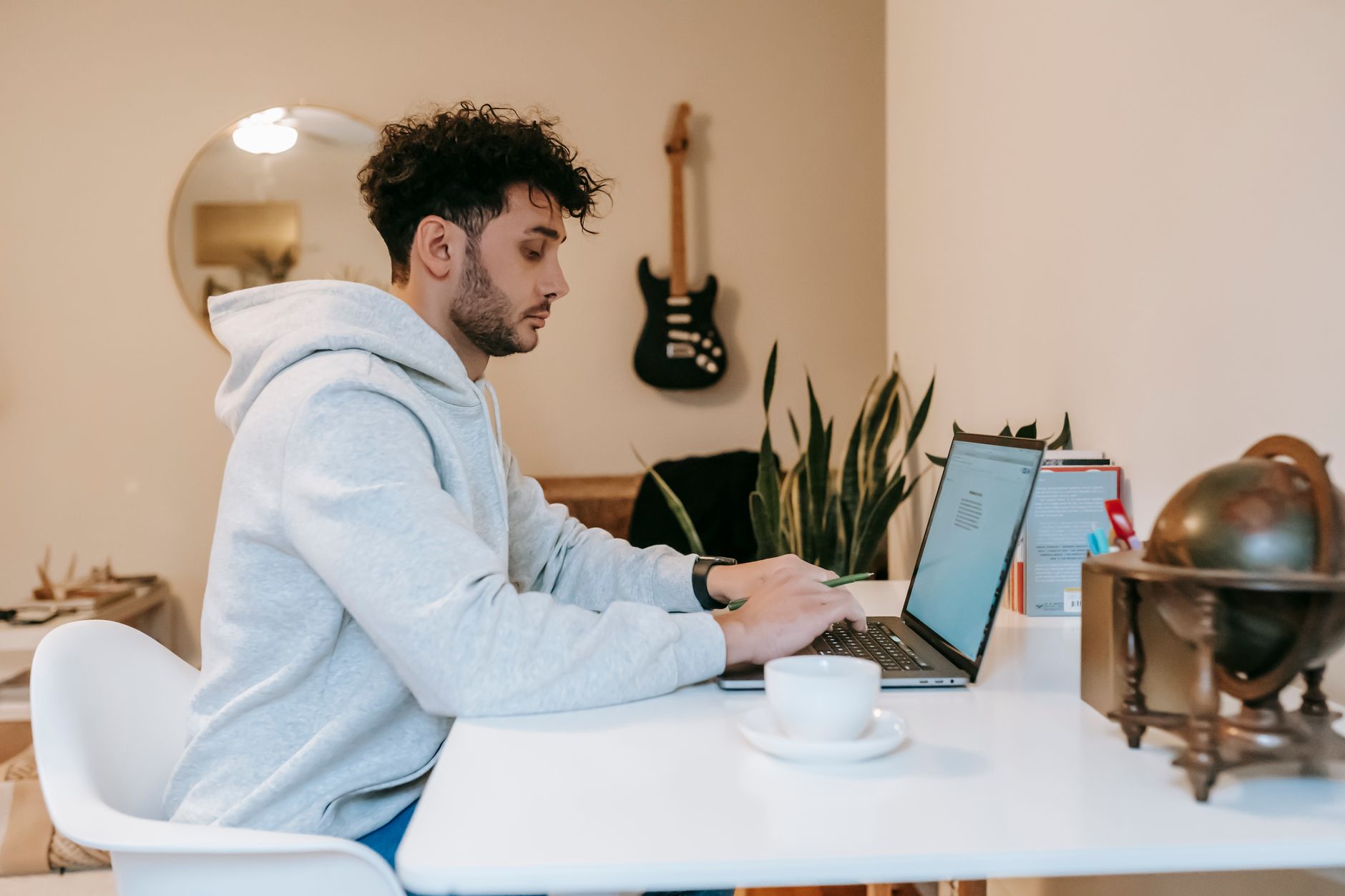 Ein Mann in grauem Kapuzenpullover sitzt an einem weissen Tisch und tippt auf einem Laptop, daneben steht eine Tasse.