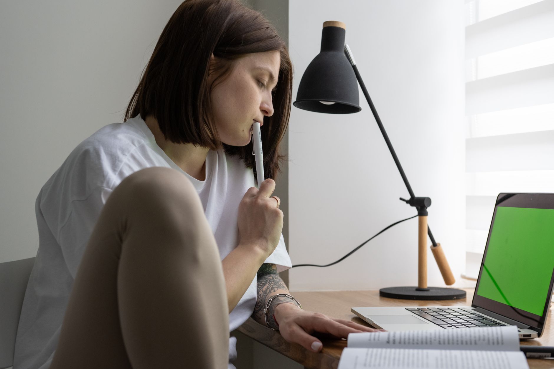 Eine junge Frau mit dunklem Bob sitzt nachdenklich am Schreibtisch, einen Stift am Mund, neben ihr eine schwarze Lampe, ein offenes Buch und ein Laptop.